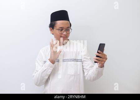 Porträt eines lächelnden jungen asiatischen muslimischen Mannes, der sich selbst fotografiert oder Videoanrufe macht, hallo sagt und seine Hand winkt. Isoliertes Bild auf weißem Hintergrund Stockfoto