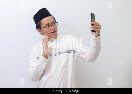 Porträt eines lächelnden jungen asiatischen muslimischen Mannes, der sich selbst fotografiert oder Videoanrufe macht, hallo sagt und seine Hand winkt. Isoliertes Bild auf weißem Hintergrund Stockfoto