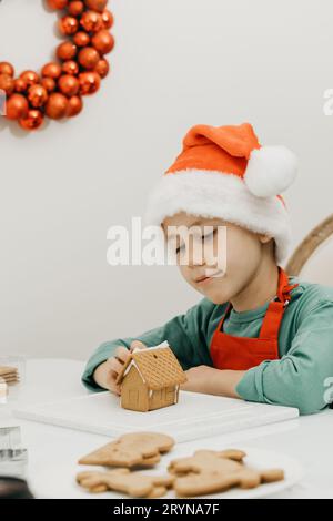 Ein süßer Junge an Silvester und Weihnachten macht in einem Weihnachtshut ein Ingwerhaus in der Küche. Stockfoto