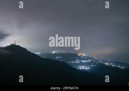 Nächtliche Aufnahme mit Nebel, der über Hügel mit einem Dorf in der Ferne rollt und unberührte Hügel von darjeeling, shimla, manali, landsdowne zeigt Stockfoto