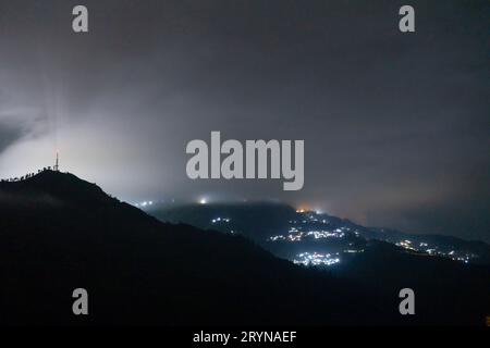 Nächtliche Aufnahme mit Nebel, der über Hügel mit einem Dorf in der Ferne rollt und unberührte Hügel von darjeeling, shimla, manali, landsdowne zeigt Stockfoto