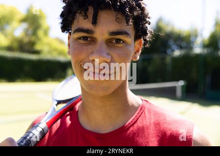 Nahaufnahme eines selbstbewussten, birassischen jungen Mannes mit lächelndem Tennisschläger auf dem Tennisplatz Stockfoto