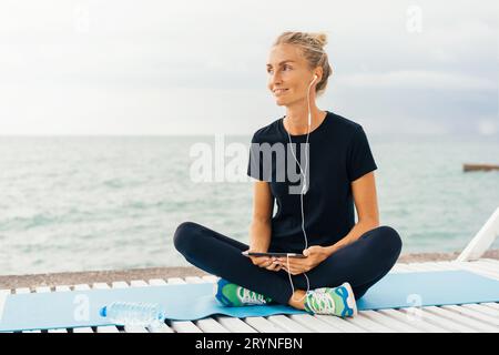 Kaukasische Frau, die sich nach dem Sport-Training am Meeresküste ausruhte und Musik oder Meditation über Kopfhörer hörte. Stockfoto