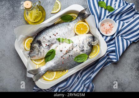 Roher Fisch dorado in Auflaufform mit Zutaten Zitrone, frischem Basilikum, Flasche Olivenöl auf hölzernem Schneidebrett mit Messer Stockfoto