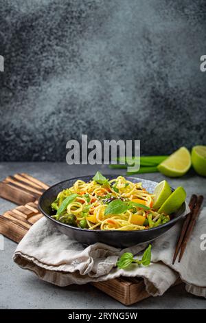 Asiatische vegetarische Nudeln mit Gemüse und Limette in schwarzer rustikaler Keramikschale, Holzstäbchen auf dem Schneidebrett, Blickwinkel auf Stockfoto