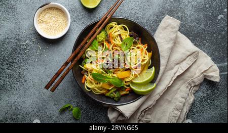 Asiatische vegetarische Nudeln mit Gemüse und Limette in schwarzer rustikaler Keramikschale, Holzstäbchen, Schneidebrett mit gehacktem Gr Stockfoto