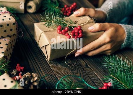 Nahaufnahme eines Weihnachtsgeschenks, eingewickelt in Bastelpapier, verziert mit Beeren und einem Fichtenzweig in weiblichen Händen. Stockfoto