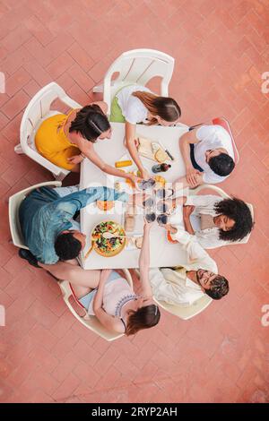 Blick auf eine Gruppe von Freunden, die ein Abendessen auf der Terrasse feiern, Rotweingläser toasten oder klirren lassen. Geselliges Treffen eines jungen erwachsenen Kumpels, der am Tisch sitzt. Hochwertige Fotos Stockfoto