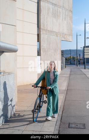 Junge, engagierte, Ingwerhaarige Frau im grünen Anzug, die mit dem Fahrrad im Finanzviertel mit modernem Gebäude im Hintergrund spazieren geht Stockfoto