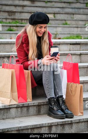 Junge kaukasische blonde Frau, die nach dem Einkaufen mit dem Smartphone ruht und im Winter auf Treppen sitzt. Stockfoto