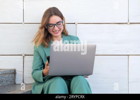 Geschäftsfrau, die auf einer Treppe vor dem Bürogebäude sitzt, während sie mit einem Laptop arbeitet Stockfoto