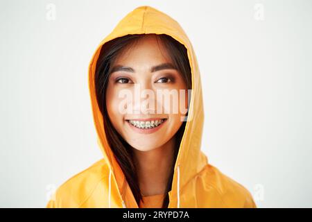 Nahaufnahme Porträt eines fröhlichen asiatischen Mädchens mit Zahnspangen in hellgelbem Regenmantel, der mit Kapuze auf dem Kopf auf weißem Studiohintergrund posiert Stockfoto