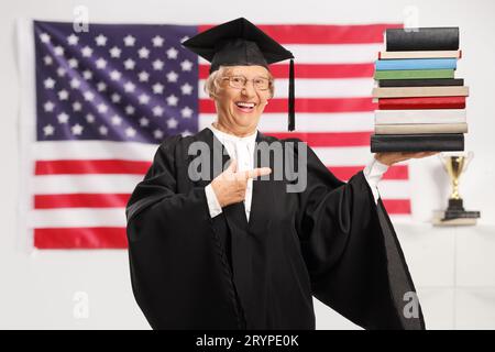 Seniorin in einem Abschlusskleid mit einem Diplom und einem Stapel Bücher mit der Flagge der USA im Hintergrund Stockfoto