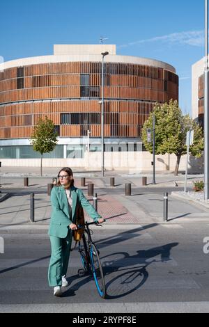 Stilvolle rothaarige Frau im grünen Anzug, die mit dem Fahrrad im Finanzviertel mit modernem Gebäude im Hintergrund spazieren geht Stockfoto