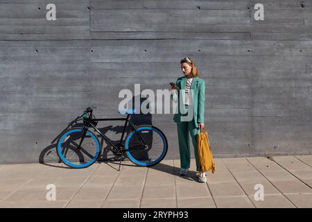 Junge, leistungsfähige, dunkelhaarige Frau im grünen Anzug, die ihr Smartphone mit ihrem Fahrrad auf schwarzem Hintergrund steht Stockfoto