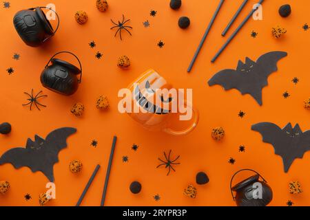 Kürbisbecher, Gummibärchen, Strohhalme, Fledermausservietten auf orangem Hintergrund. Halloween-Feier-Konzept Stockfoto