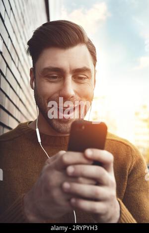 Porträt eines lächelnden Mannes mit Kopfhörern und Handy, das an einer Ziegelwand steht. Stockfoto
