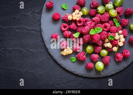 Schieferschale mit verschiedenen Beeren. Himbeeren, Stachelbeeren, Johannisbeeren auf schwarzem Hintergrund Stockfoto