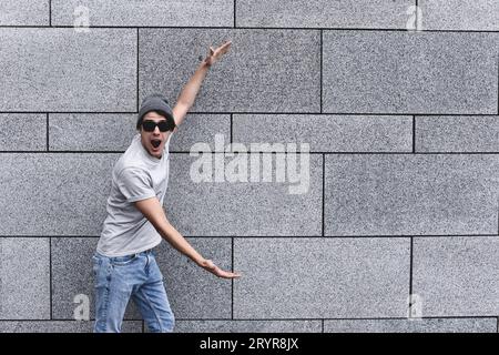 Hübscher junger Mann mit Sonnenbrille, der mit schockiertem Gesichtsausdruck in die Kamera blickt und vor grauer Wand steht. Stockfoto