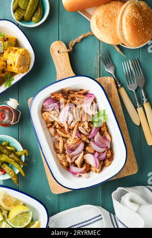 Gezapftes bbq-Hühnchen mit gebackenen Zwiebeln auf Servierplatte, bbq-Mais, Gurken, Chilischoten und Brötchen für Hot Dogs und Burger, Tomatensauce. Traditionell Stockfoto