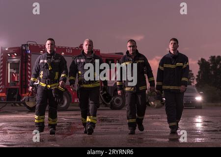 Mutiges Feuerwehrteam geht zur Kamera. Im Hintergrund kämpfen Sanitäter und Feuerwehrleute Rettungsdienst Feuer bei Autounfall, INSU Stockfoto