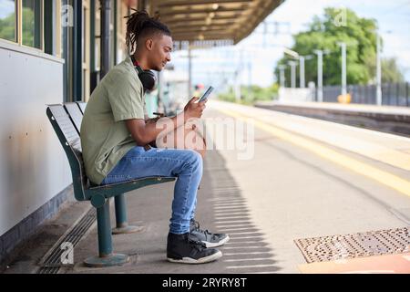 Junger Mann, Der Im Zug Zur Arbeit Pendelt, Sitzt Auf Der Plattform Und Schaut Auf Ein Mobiltelefon Für Reiseinformationen Oder Soziale Medien Stockfoto