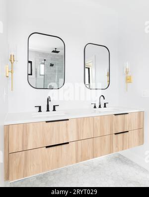 Badezimmer mit einem Schrank aus Naturholz, weißer Marmorarbeitsplatte mit schwarzen Armaturen, schwarz gerahmten Spiegeln und goldenen Wandleuchten. Stockfoto