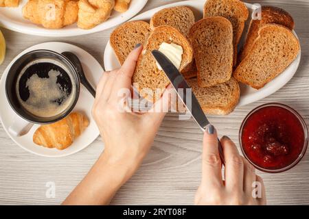 Weibliche Hände verteilen Butter auf einen Toast mit einer Tasse schwarzen Kaffee Stockfoto