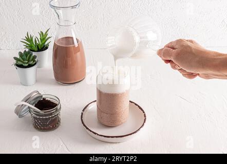 Die Hand einer Frau gießt geschlagene Milch aus einer Glaskanne in ein modisches Glas in Form einer Bierdose in Kaffee. Kaffee Latte, Cappuccino. Stockfoto