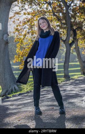 Eine Frau in gemütlicher Herbstkleidung steht inmitten des bunten Laubs großer Bäume, die mit Herbstlaub geschmückt sind Stockfoto