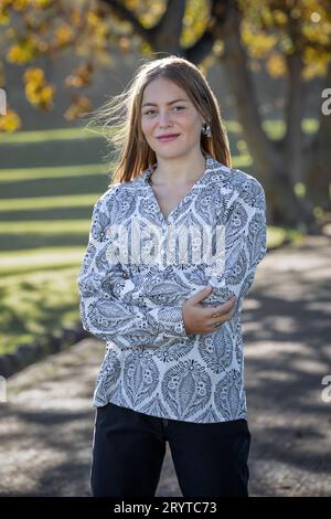 In einer Nahaufnahme steht eine junge blonde Frau unter goldenen Herbstbäumen, elegant gekleidet in einem schwarz-weiß gemusterten Hemd Stockfoto