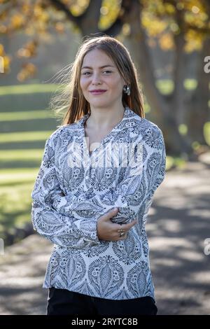 In einer Nahaufnahme steht eine junge blonde Frau unter goldenen Herbstbäumen, elegant gekleidet in einem schwarz-weiß gemusterten Hemd Stockfoto