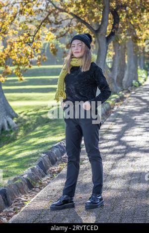 Eine junge blonde Frau in gemütlicher schwarzer Kleidung steht anmutig unter goldenen Herbstbäumen Stockfoto