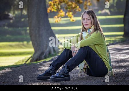 Eine sitzende junge blonde Frau in gemütlicher Kleidung sitzt unter Herbstbäumen, ihre Arme liegen lässig auf den Knien Stockfoto