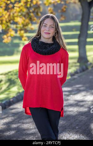 In einem Kopf- und Schulterschuss strahlt eine atemberaubende junge blonde Frau, die einen schwarzen Snood und einen knallroten Pullover trägt, unter den Herbstbäumen aus Stockfoto