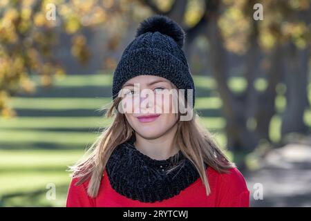 In einem Kopf- und Schulterschuss strahlt eine atemberaubende junge blonde Frau, die einen schwarzen Wollmütze und einen knallroten Pullover trägt, unter den Herbstbäumen aus Stockfoto