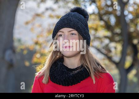 In einem Kopf- und Schulterschuss strahlt eine atemberaubende junge blonde Frau, die einen schwarzen Wollmütze und einen knallroten Pullover trägt, unter den Herbstbäumen aus Stockfoto