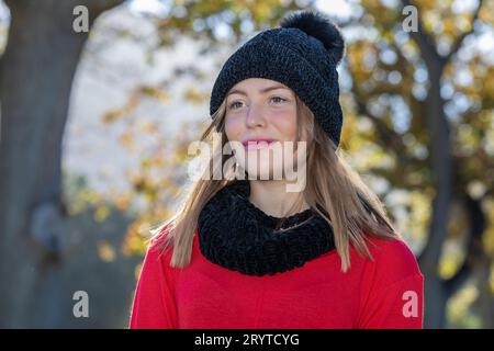 In einem Kopf- und Schulterschuss strahlt eine atemberaubende junge blonde Frau, die einen schwarzen Wollmütze und einen knallroten Pullover trägt, unter den Herbstbäumen aus Stockfoto