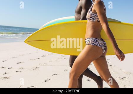 Ein afroamerikaner-Paar mit Surfbrettern, das auf dem sonnigen Strand zum Meer spaziert Stockfoto