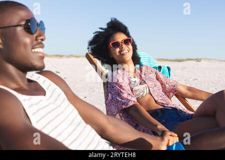 Glückliches afroamerikanisches Paar, das auf Liegestühlen sitzt und sich am sonnigen Strand die Hände hält Stockfoto