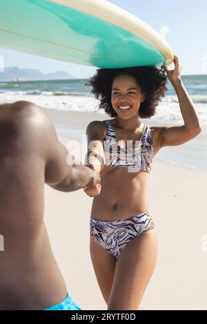 Glückliches afroamerikanisches Paar, das ein Surfbrett auf den Köpfen hält, Hände am sonnigen Strand hält Stockfoto
