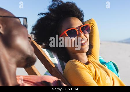 Glückliches afroamerikanisches Paar mit Sonnenbrille, das auf Liegestühlen sitzt und am sonnigen Strand lächelt Stockfoto