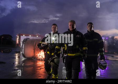 Mutiges Feuerwehrteam geht zur Kamera. Im Hintergrund kämpfen Sanitäter und Feuerwehrleute Rettungsdienst Feuer bei Autounfall, INSU Stockfoto