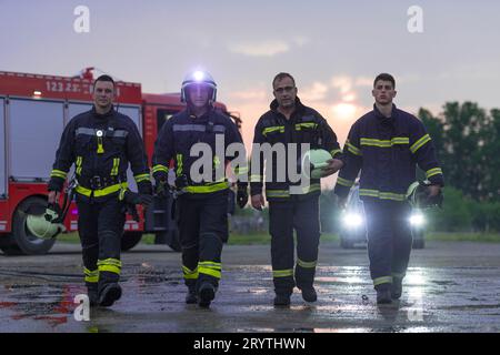 Mutiges Feuerwehrteam geht zur Kamera. Im Hintergrund kämpfen Sanitäter und Feuerwehrleute Rettungsdienst Feuer bei Autounfall, INSU Stockfoto