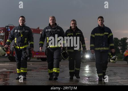 Mutiges Feuerwehrteam geht zur Kamera. Im Hintergrund kämpfen Sanitäter und Feuerwehrleute Rettungsdienst Feuer bei Autounfall, INSU Stockfoto