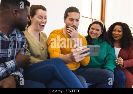 Glückliche, vielfältige Freunde, die sich zu Hause entspannen, miteinander reden und auf das Smartphone schauen Stockfoto