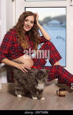 Eine schwangere Frau im Wohnzimmer mit einer Tasse Tee sitzt auf der Schwelle einer Glastür und spielt mit der Katze. Stockfoto