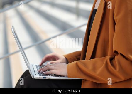 Nahaufnahme einer jungen Freelancerin im braunen Blazer, die Tasten der Laptop-Tastatur berührt, während sie an Projekten im städtischen Umfeld arbeitet Stockfoto