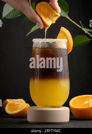 Eiskaffee mit Orangensaft in einem durchsichtigen Glas, die Hand einer Frau drückt eine Orangenscheibe in ein Seifenblasengetränk Stockfoto