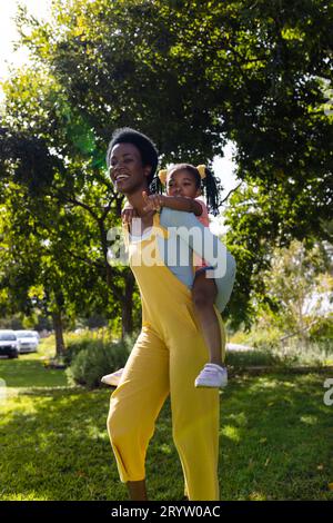 afroamerikanische Mutter, die sich mit dem Huckepack beschäftigt, fröhliche Tochter, während sie auf dem grasbewachsenen Feld im Park spaziert Stockfoto